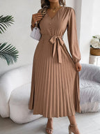 Woman in a brown long-sleeve V-neck midi dress with a pleated skirt and a self-tie belt, carrying a white handbag