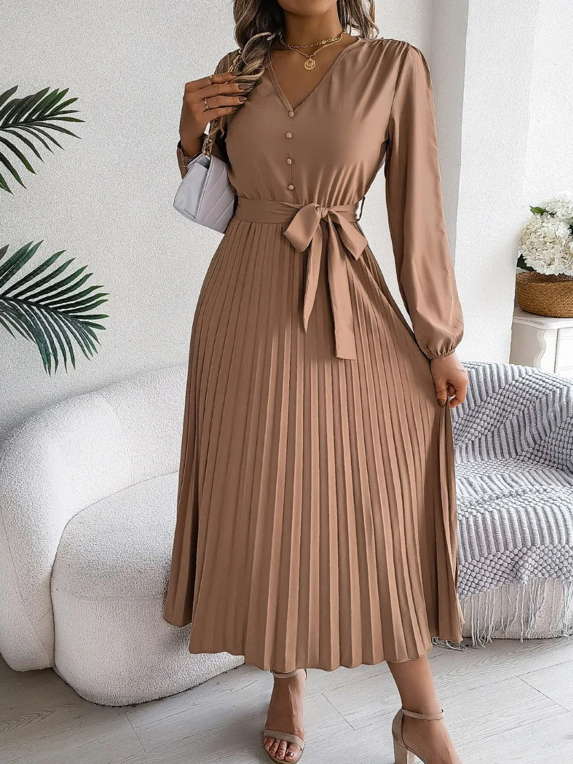 Woman in a brown long-sleeve V-neck midi dress with a pleated skirt and a self-tie belt, carrying a white handbag