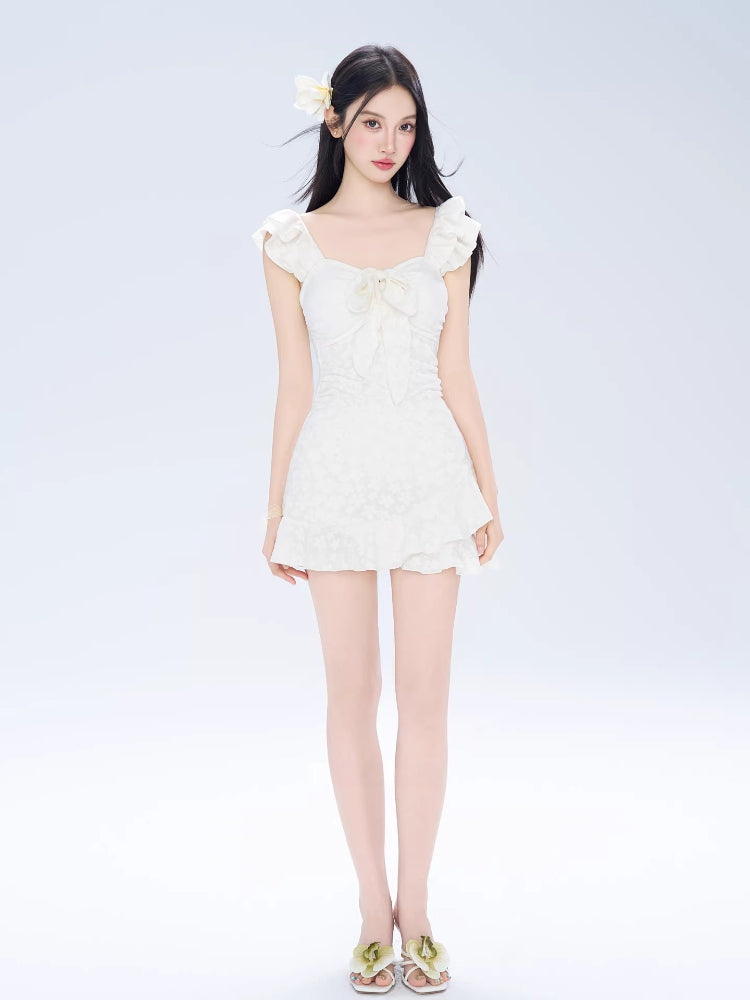 Woman wearing a white jacquard fit and flare mini dress with ruffled cap sleeves and a sweetheart neckline with a bow detail.