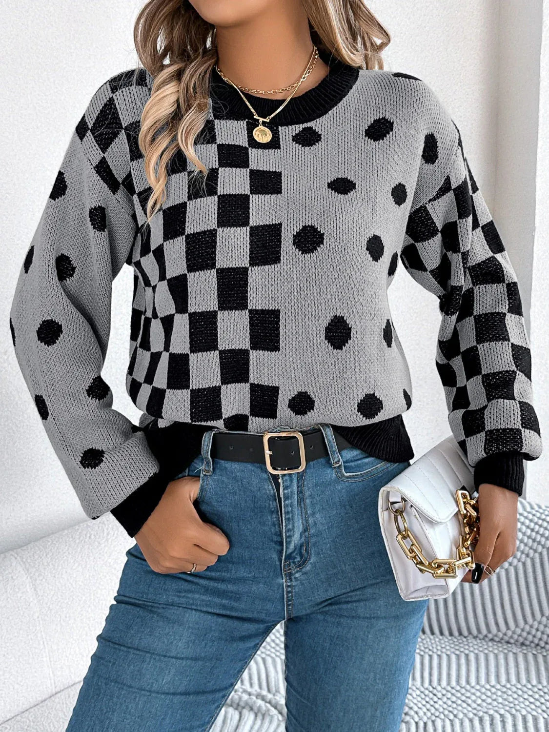 Woman wearing a grey knit sweater with a black checkerboard and polka dot pattern, paired with blue jeans and a white handbag.