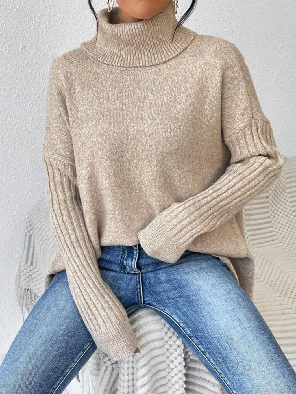 Woman sitting and wearing a beige knit turtleneck tunic with ribbed sleeves and asymmetrical hem.