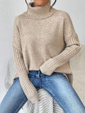Woman sitting and wearing a beige knit turtleneck tunic with ribbed sleeves and asymmetrical hem.