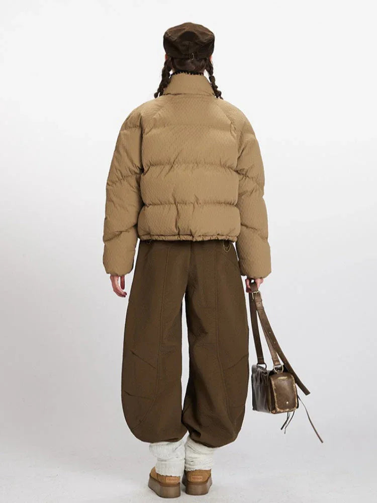 Back view of a tan quilted puffer jacket with a high collar and black trim, worn with olive green wide-leg pants and a brown cap