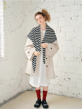 Full-body shot of a woman wearing a black and white striped scarf, beige coat, white dress, red socks, and black shoes.