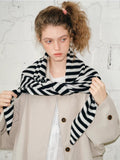 Woman holding the ends of a black and white striped scarf, styled over a beige coat.