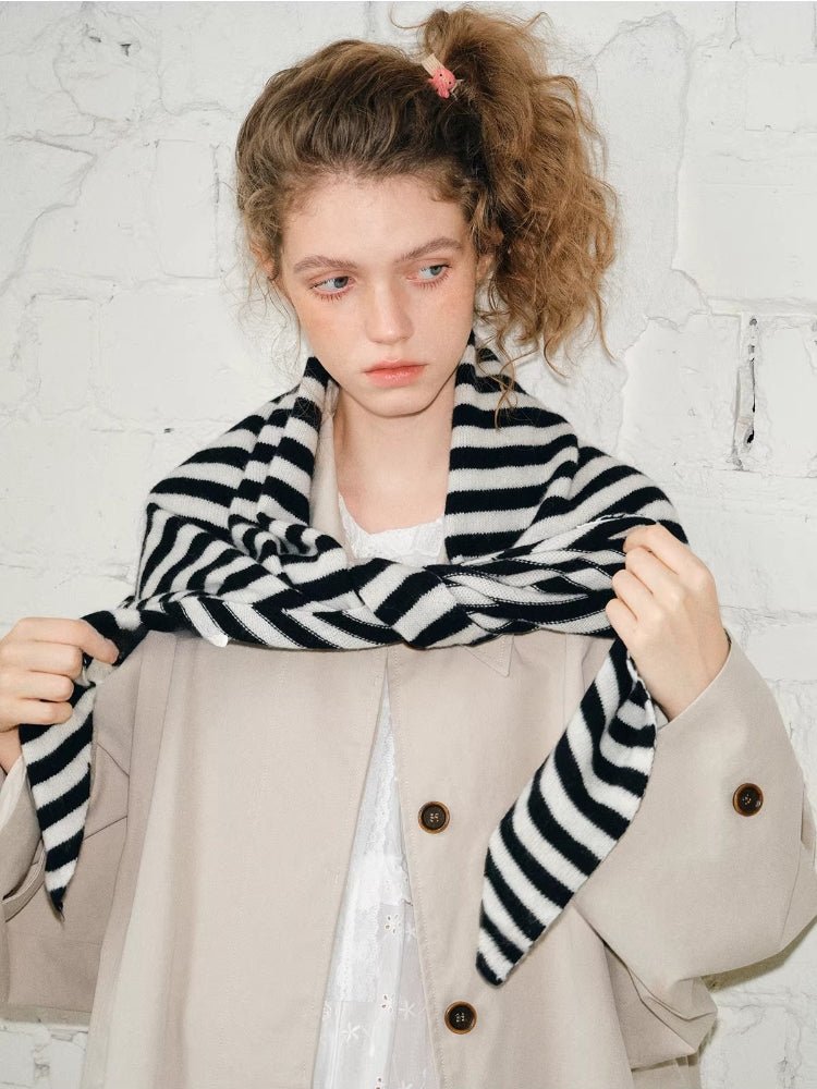 Woman holding the ends of a black and white striped scarf, styled over a beige coat.