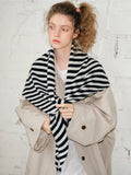 Woman tying a black and white striped scarf at the front, layered over a beige coat.