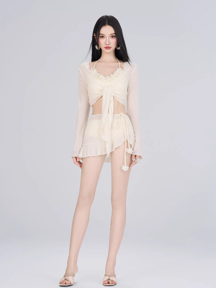 Full body shot of a woman wearing a cream-colored three-piece beachwear set including a sheer cover-up, bikini top, and wrap skirt.