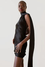 woman wearing a black satin mini dress with a high neckline and a long sash, shown from the side