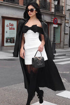 Woman in a white and black dress, black trench coat, and accessories walking on a city street.