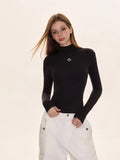 Woman wearing a black turtleneck top with a silver embroidered cross logo on the chest.