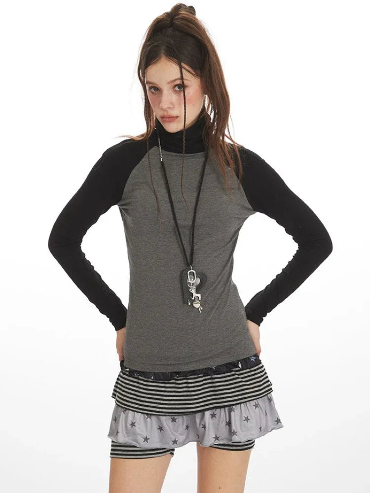 Close-up front view of a woman wearing a grey long-sleeved turtleneck top with black raglan sleeves and a pendant necklace.