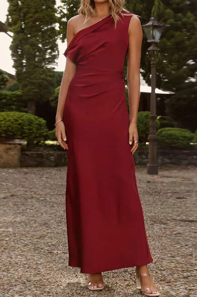 Woman standing in a garden wearing a long burgundy satin dress with a single shoulder strap.