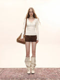 Woman standing on a rug wearing a cream long-sleeve top, brown shorts, and cream slouchy boots, with a shoulder bag.