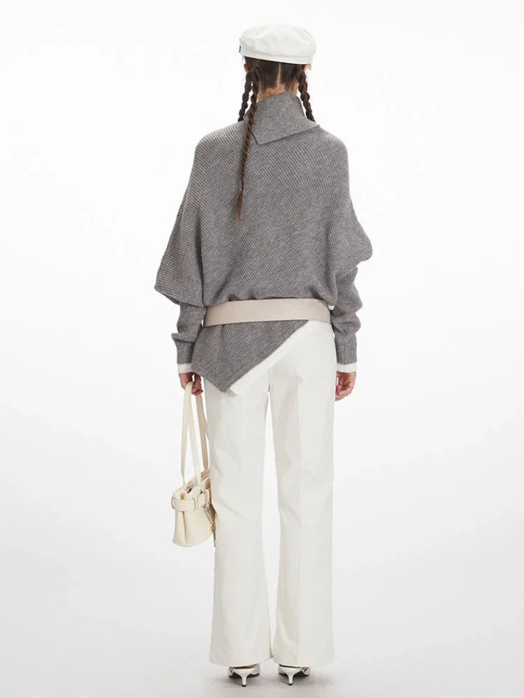 Woman wearing a grey knit poncho with a turtleneck and batwing sleeves, white pants, a cream belt, and a white handbag.