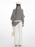 Back view of a woman wearing a grey knit poncho with a turtleneck and batwing sleeves, white pants, a cream belt, and a white handbag.