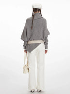 Back view of a woman wearing a grey knit poncho with a turtleneck and batwing sleeves, white pants, a cream belt, and a white handbag.