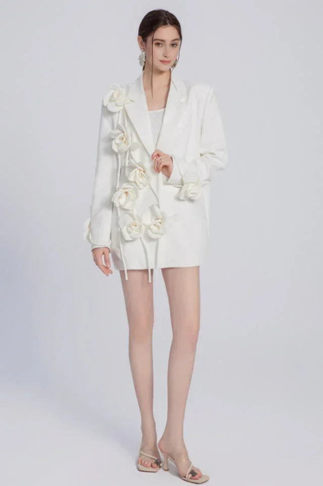 A woman models a white blazer adorned with fabric flowers, presented against a plain background.