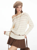 Close-up of a model wearing a cream-colored off-the-shoulder knit sweater with a diamond pattern and decorative trim. The sweater is styled with a decorative belt and a brown textured mini skirt.