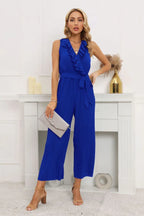 Woman wearing a royal blue sleeveless pleated jumpsuit with a ruffled V-neck and a tie belt.