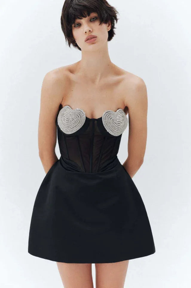 Model posing in a black strapless mini dress with a corset bodice adorned with heart-shaped embellishments and a sheer midsection, against a white background.