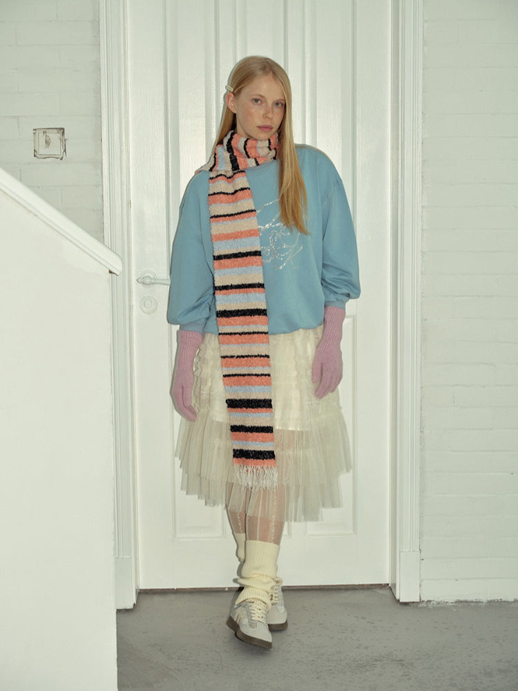 Woman wearing a light blue sweatshirt, a white tulle skirt, pink gloves, white socks, and sneakers, with a multi-colored striped scarf around her neck.