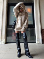 Woman styled in a beige oversized knit hooded tunic with a black handbag and sunglasses.