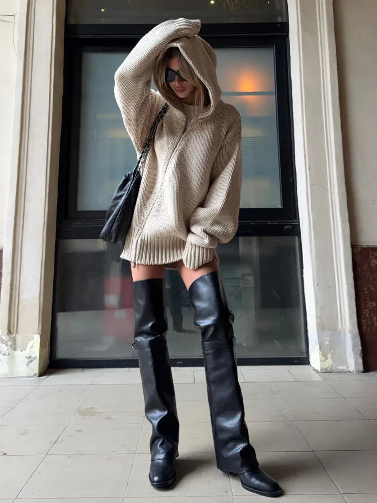 Woman styled in a beige oversized knit hooded tunic with a black handbag and sunglasses.