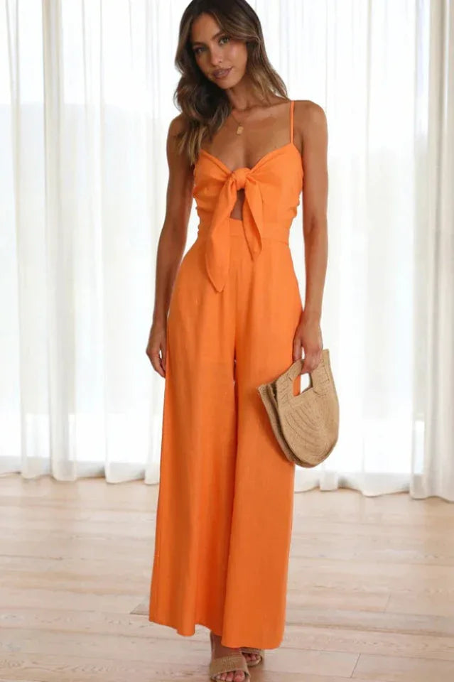 Woman wearing an orange wide leg jumpsuit with a tie-front detail and spaghetti straps, standing indoors. Main image