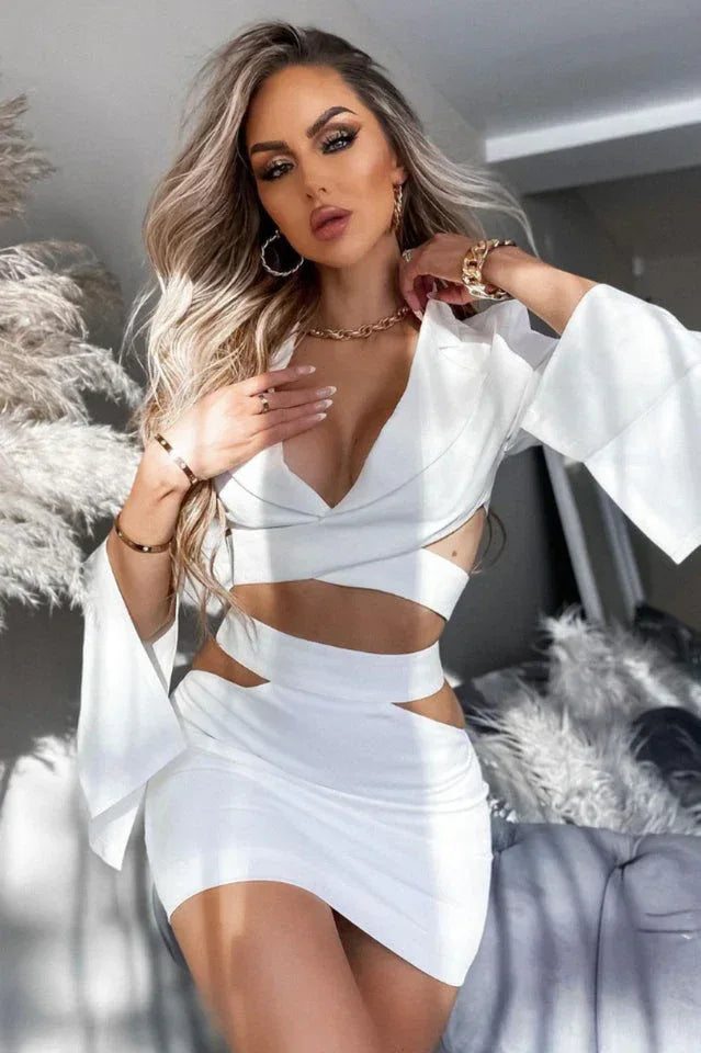 Woman with blonde hair wearing a white long-sleeve crop top and mini skirt with cut-out details, accessorized with jewelry.