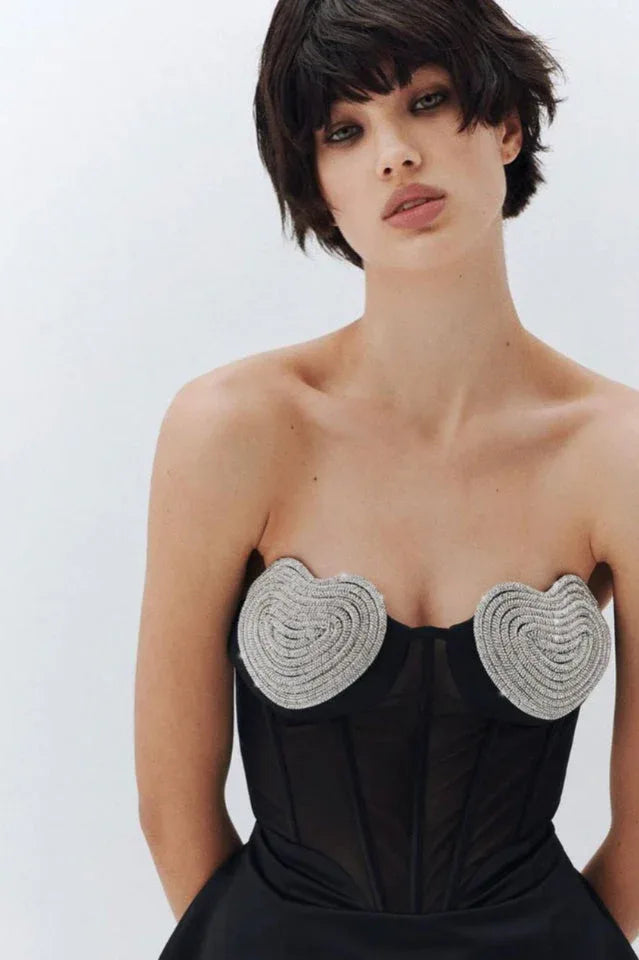 Close-up of a model's torso wearing a black strapless mini dress with a detailed corset bodice featuring heart-shaped embellishments.