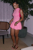 Woman wearing a pink sleeveless ribbed mock neck mini dress with a bubble hem skirt and gold wedge sandals, standing indoors.