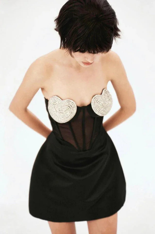 A model in a black strapless mini dress with a corset bodice and heart-shaped embellishments, looking down against a white backdrop.