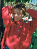Woman wearing a coral knit sweater with her hands in her hair, showcasing the button detail and ribbed texture.