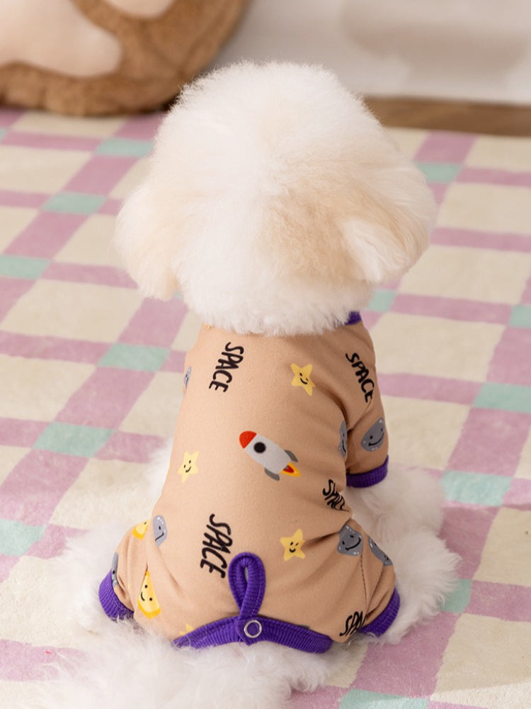 Small white dog wearing a tan dog pajama with a space print and purple trim