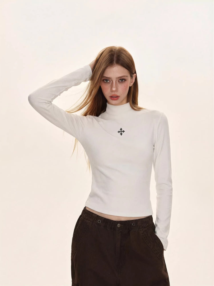 Woman wearing a white turtleneck top with a black embroidered cross logo on the chest.