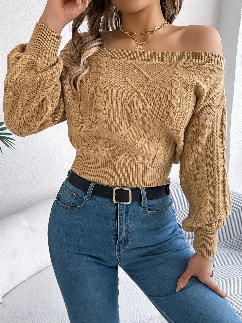 Woman wearing a tan off-the-shoulder cable knit cropped sweater with balloon sleeves and blue jeans.