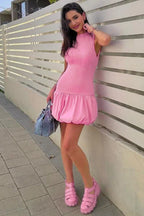 Woman wearing a pink sleeveless ribbed mock neck mini dress with a bubble hem skirt and pink platform sandals, posing outdoors.