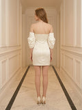 Back view of a woman in a cream off-the-shoulder satin mini dress with puff sleeves, standing in a hallway.