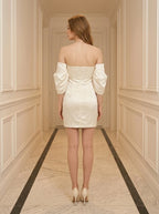 Back view of a woman in a cream off-the-shoulder satin mini dress with puff sleeves, standing in a hallway.