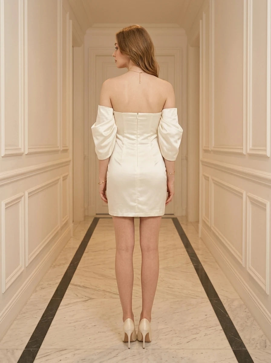 Back view of a woman in a cream off-the-shoulder satin mini dress with puff sleeves, standing in a hallway.