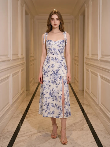 Woman wearing a white midi dress with a blue floral print and tie shoulder straps, standing in a hallway.