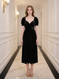 Woman wearing a black floral burnout velvet midi dress with puff sleeves and a sweetheart neckline, standing in a hallway.