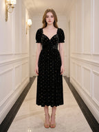 Woman wearing a black floral burnout velvet midi dress with puff sleeves and a sweetheart neckline, standing in a hallway.