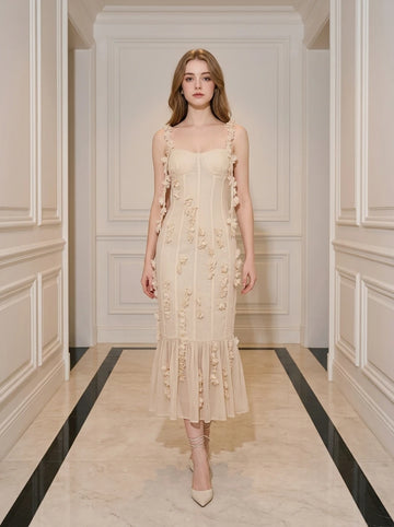 Woman wearing a cream-colored midi dress with 3D floral embellishments and a ruffled hem, standing in a hallway.