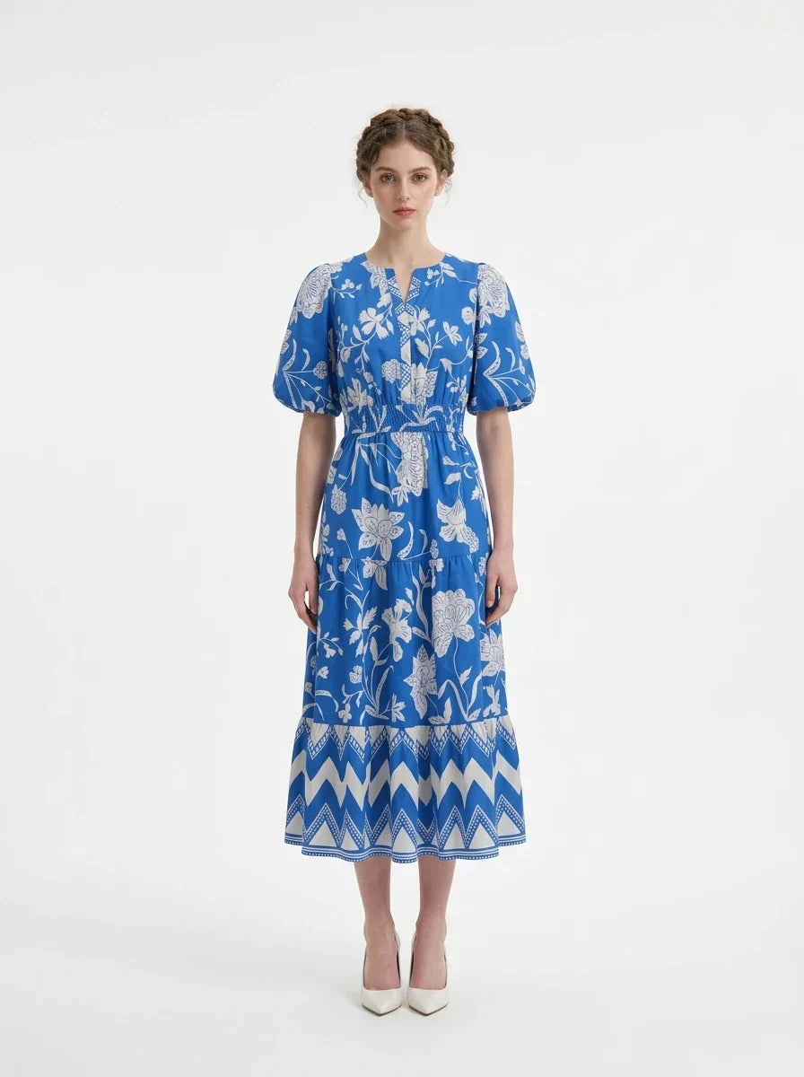 Woman wearing a blue floral print midi dress with puff sleeves and a tiered skirt, standing against a white background.