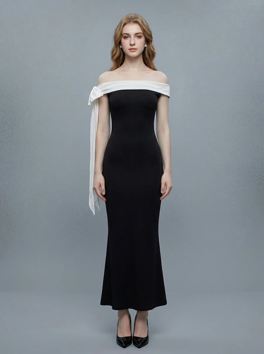 Woman in a black off-the-shoulder mermaid dress with a white draped accent on the shoulder, standing against a grey background.