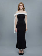 Woman in a black off-the-shoulder mermaid dress with a white draped accent on the shoulder, standing against a grey background.