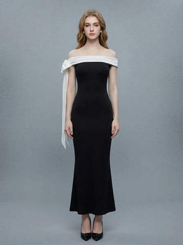 Woman in a black off-the-shoulder mermaid dress with a white draped accent on the shoulder, standing against a grey background.