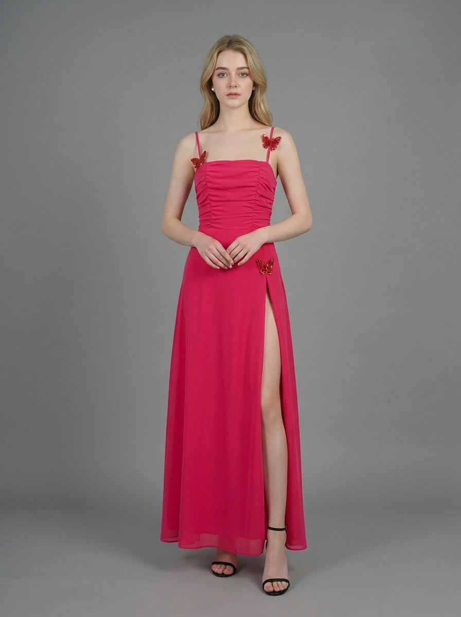 Woman in a bright pink ruched maxi dress with spaghetti straps and butterfly details, standing against a grey backdrop.